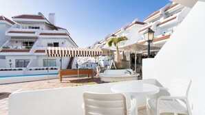 Un espace aménagé au bord de la piscine, avec des chaises blanches et une table, une piscine et un bâtiment doté de plusieurs balcons.