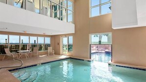 An indoor swimming pool with clear blue water, surrounded by large windows offering views of the outside.