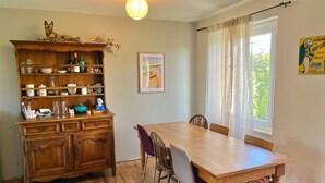 A dining area with a wooden table and chairs, a wooden cabinet with glassware, and a window with curtains. - L'Authentik (Gouesnach)