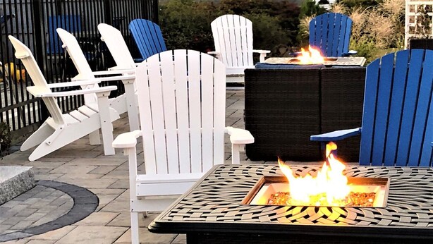 Een patio met witte en blauwe Adirondack-stoelen, een vierkant vuurkorf met vlammen en een geweven tafel.