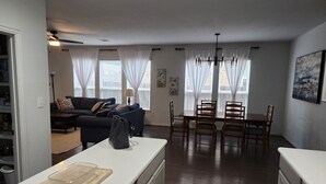 A modern kitchen with a dining area, a living room with a sofa and a coffee table, and large windows with white curtains.
