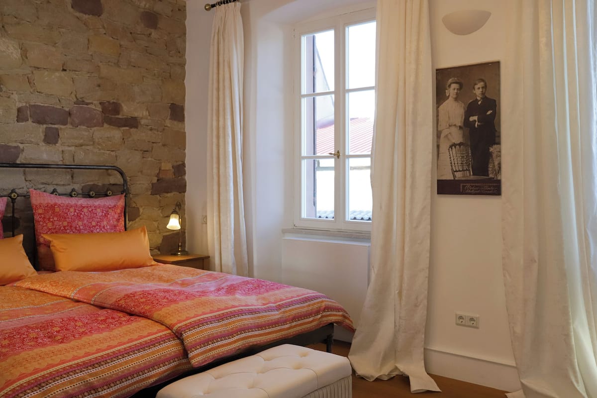 A bedroom with a stone wall, a bed with a patterned bedspread, a nightstand with a lamp, and a window with curtains.