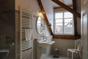 Une salle de bain avec un lavabo blanc, un miroir, un porte-serviettes et une fenêtre donnant sur des immeubles.
