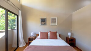 Un dormitorio con una cama grande, dos mesitas de noche con lámparas, una ventana con vistas a zonas verdes y dos cuadros en la pared.