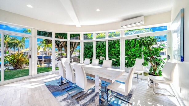 A modern dining area with a large table and chairs, large windows with a view of greenery and palm trees, and a ceiling with recessed lighting. - Heated Pool + Waterfront! Close 2 Beach & Shops! (Pompano Beach)