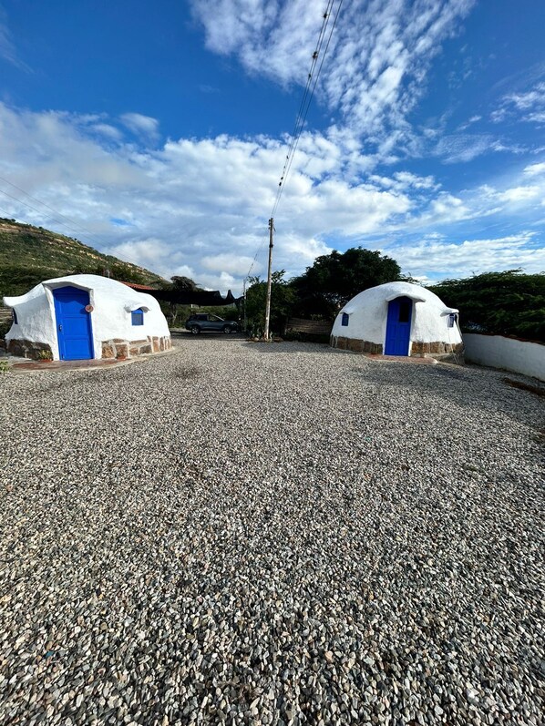 Two white yurts with blue doors on a gravel surface, surrounded by greenery and a clear sky.