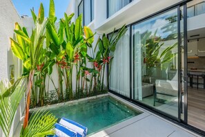 Un área de piscina moderna al aire libre con un porche de cristal, rodeada de exuberante follaje tropical y una piscina de aguas cristalinas.