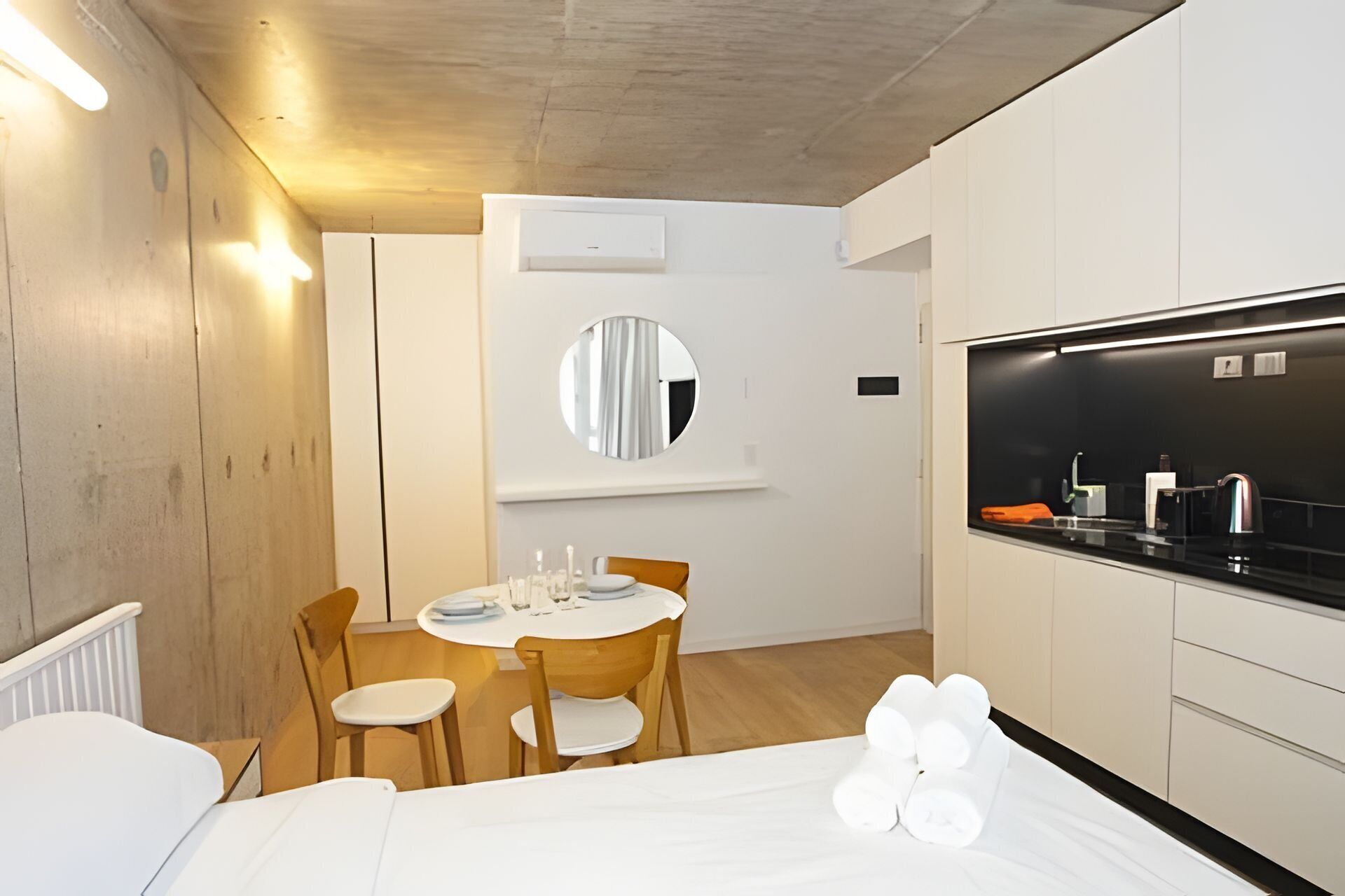 A modern hotel room with a wooden floor, a round dining table with chairs, a bed with white linens, a large mirror, and a black-framed TV.