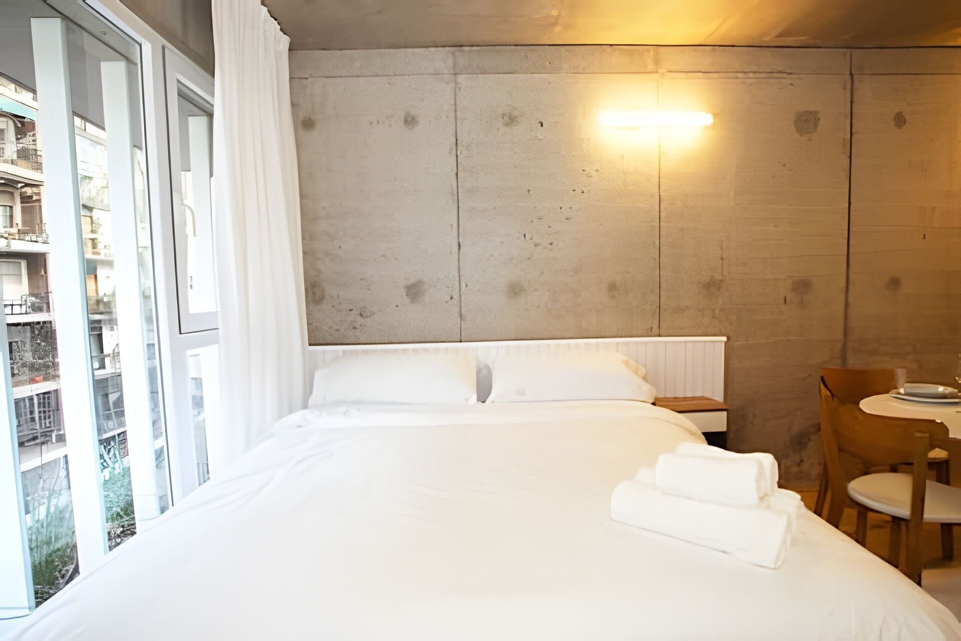 A hotel room with a large bed, a bedside table, a chair, and a view of the city through large windows.