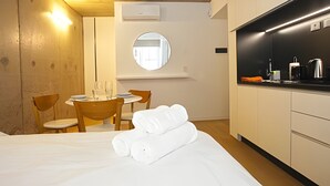 Quarto de hotel compacto com cama, uma pequena mesa de jantar com cadeiras, um espelho redondo e uma cozinha compacta.