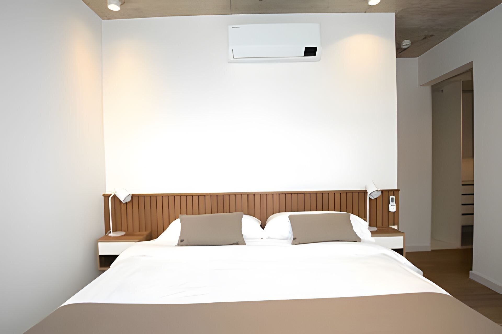 A modern hotel room with a large bed, wooden headboard, bedside tables with lamps, and a built-in air conditioning unit.