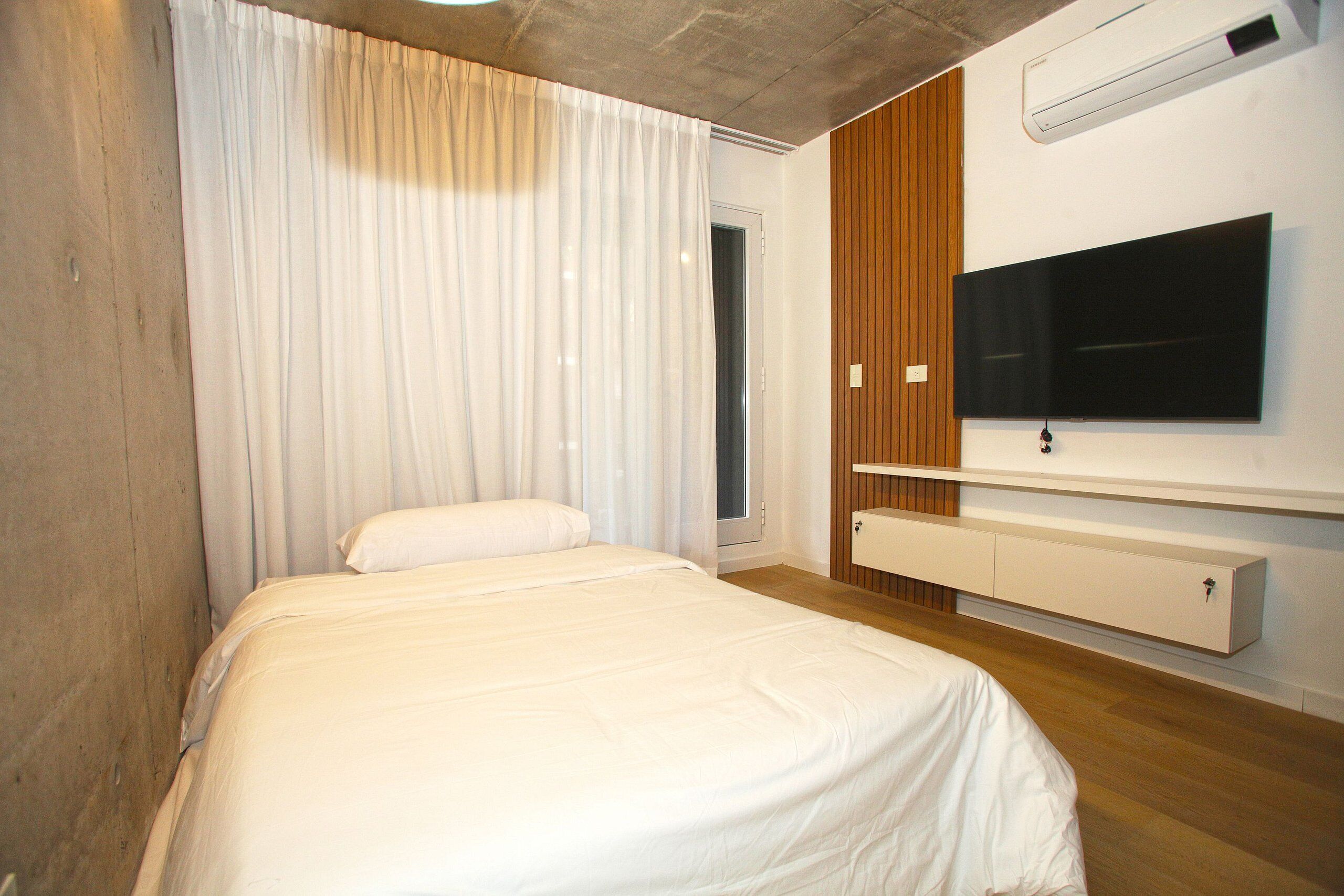 Un dormitorio con una cama, un televisor instalado en la pared y un ventanal con cortinas.