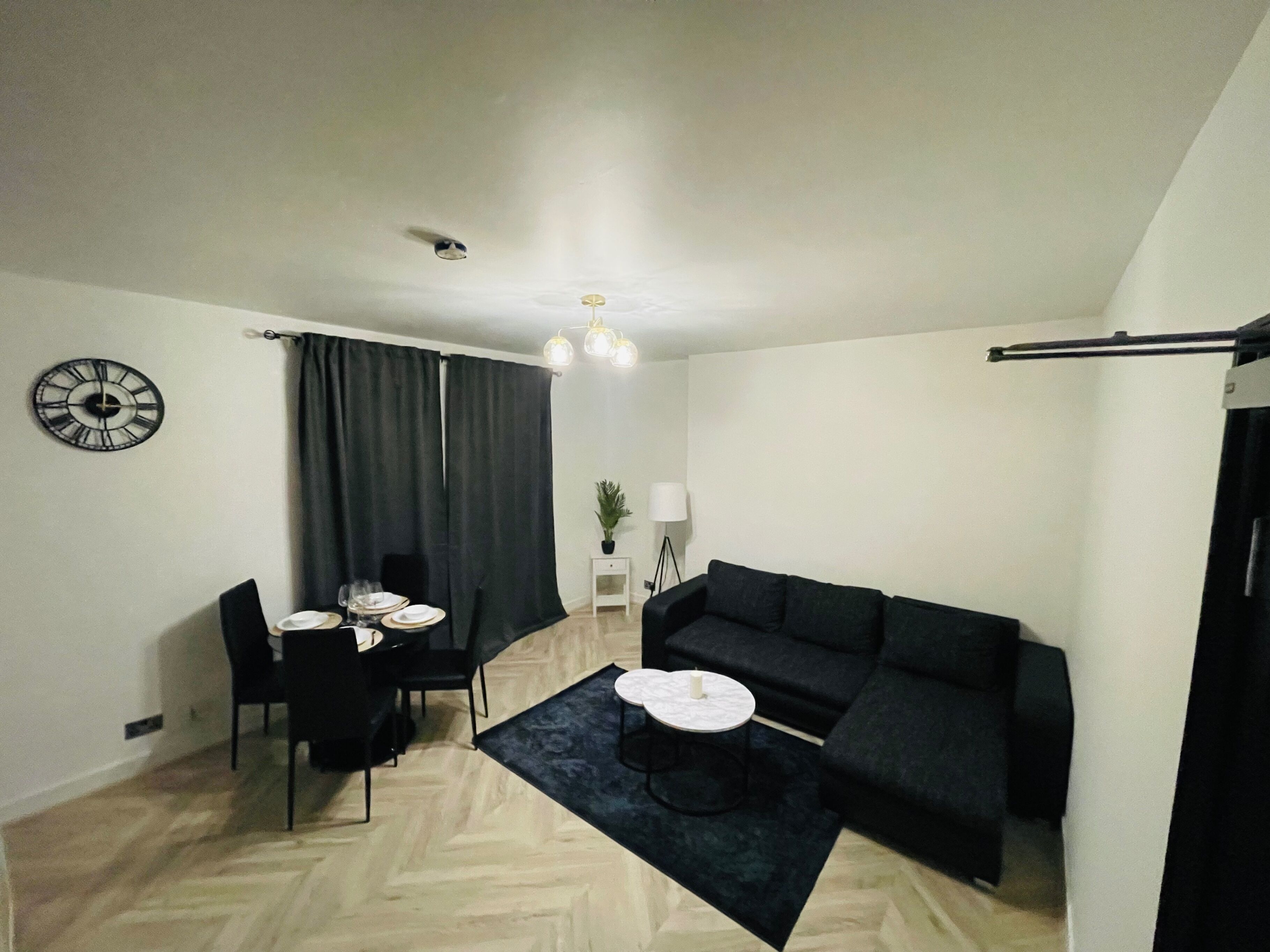 Una sala de estar moderna con un sofá negro, una mesa de comedor con sillas, una mesita auxiliar, un reloj en la pared y una lámpara en el techo.