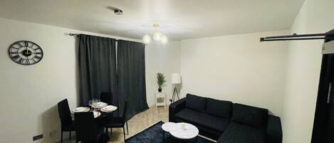 Una sala de estar moderna con un sofá negro, una mesa de comedor con sillas, una mesita auxiliar, un reloj en la pared y una lámpara en el techo.