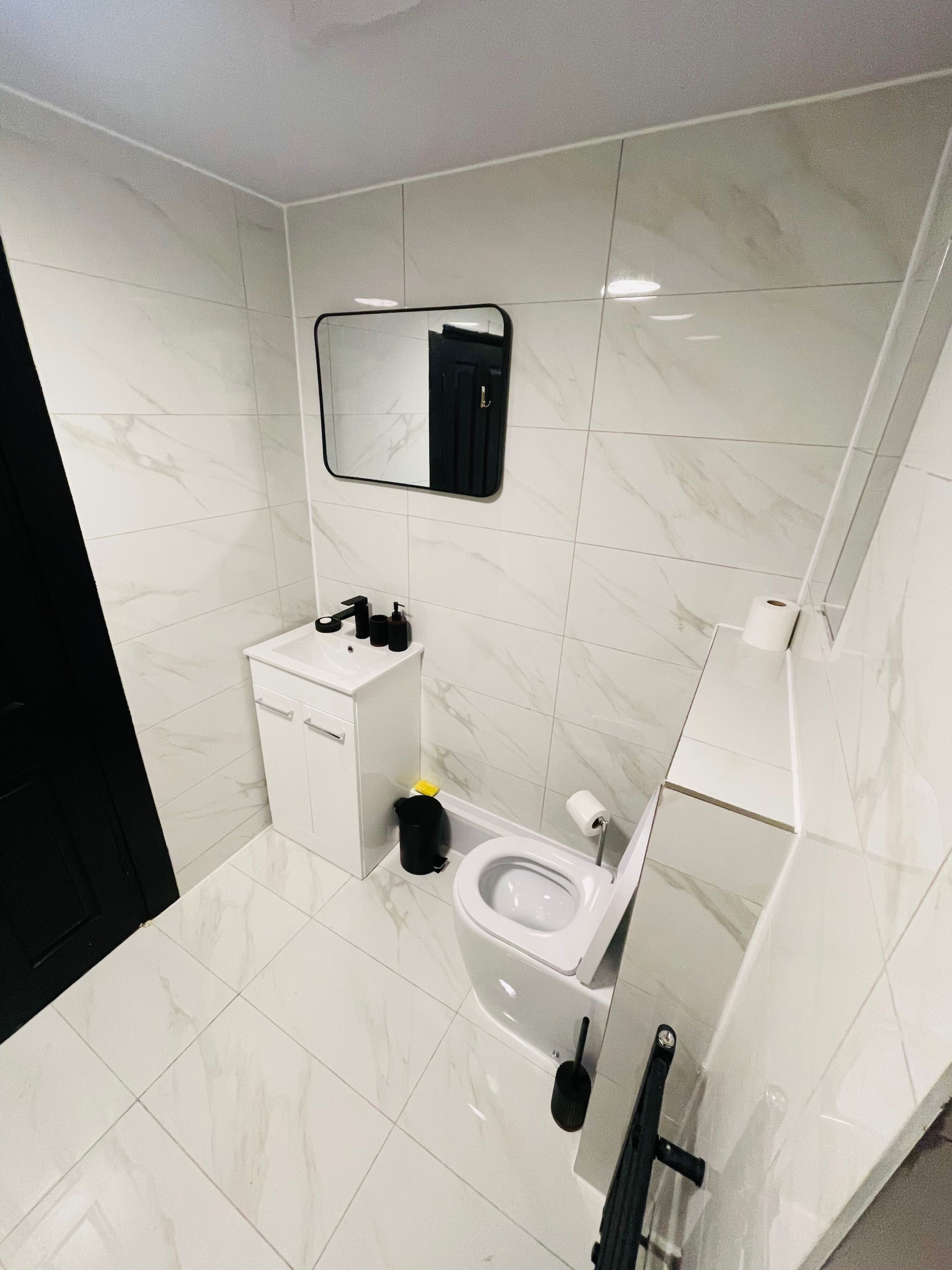 A modern bathroom with white marble tiles, a toilet, a sink with a mirror above, and a black trash bin.