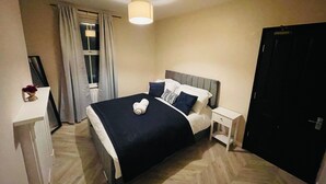 Une chambre à coucher comprenant un lit, des tables de chevet, une commode, une fenêtre avec des rideaux, une porte et un luminaire au plafond.