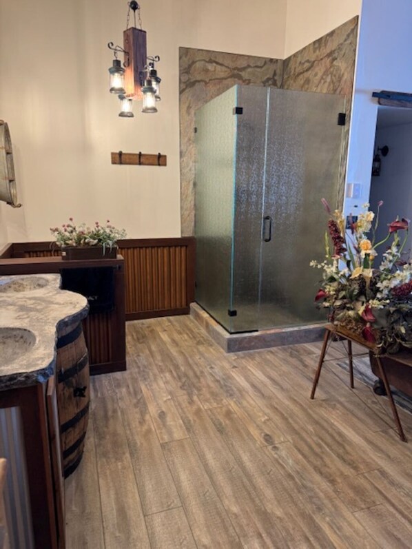 A bathroom with a glass shower enclosure, wooden flooring, a wooden bench, and a small flower arrangement. - Paradise at Legacy Ranch (Twin Lakes)