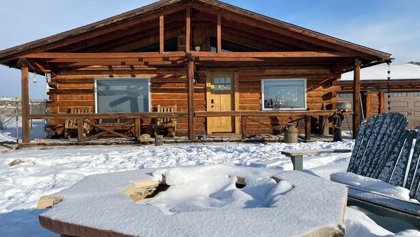 Una cabaña de madera con un porche, cimientos de piedra y una zona de estar exterior cubierta de nieve.