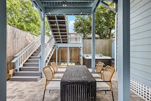 A covered patio with a wooden table and chairs, a hot tub, and a staircase leading to a deck. - The Lookout at R½ | Hot Tub • Sauna • Wraparound Deck • Steps to Beach (Galveston)