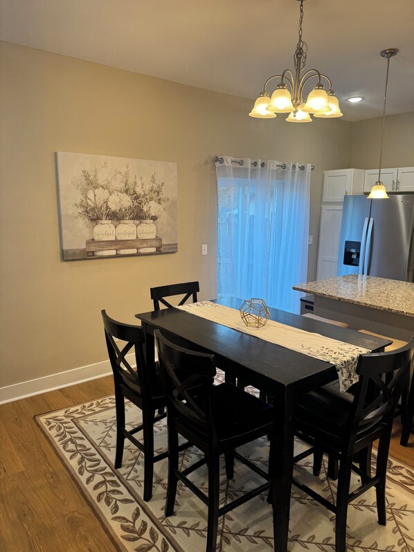 A dining area with a black table and chairs, a chandelier, a refrigerator, and a window with curtains.
