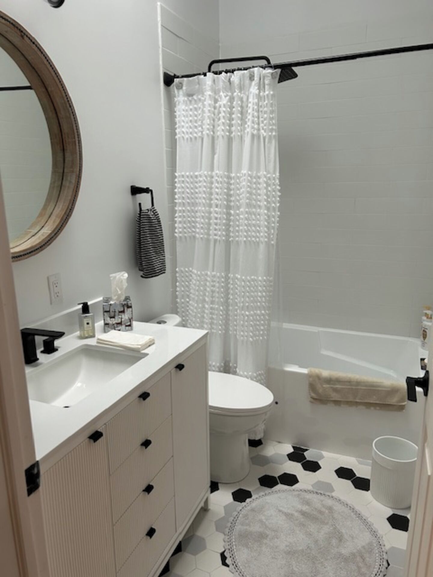 Un bagno moderno con lavandino, water e vasca da bagno bianchi. Sopra il lavandino c'è uno specchio rotondo, un asciugamano a righe appeso al muro e una tenda da doccia bianca. Il pavimento presenta un motivo a esagoni bianco e nero.