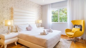 Une chambre d’hôtel avec un grand lit, deux tables de chevet avec des lampes, un fauteuil jaune et une fenêtre donnant sur un paysage verdoyant.