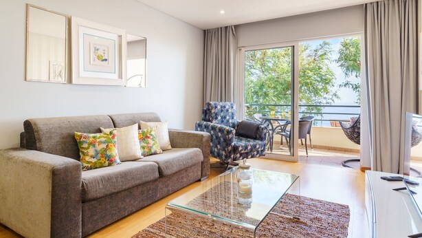 Un salon comprenant un canapé, un fauteuil et une table basse en verre. Il y a également un balcon avec du mobilier d’extérieur.