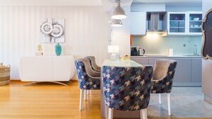 Un salon moderne avec un canapé blanc, des fauteuils à motifs, une table basse en verre et une table à manger. On aperçoit, au fond, un coin cuisine avec des placards et un évier.