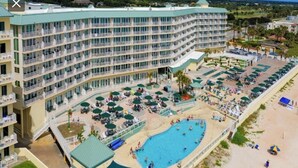 Ein großes Hotel mit zahlreichen Balkonen, einem zentral gelegenen Pool und einer Strandbucht mit Liegestühlen.