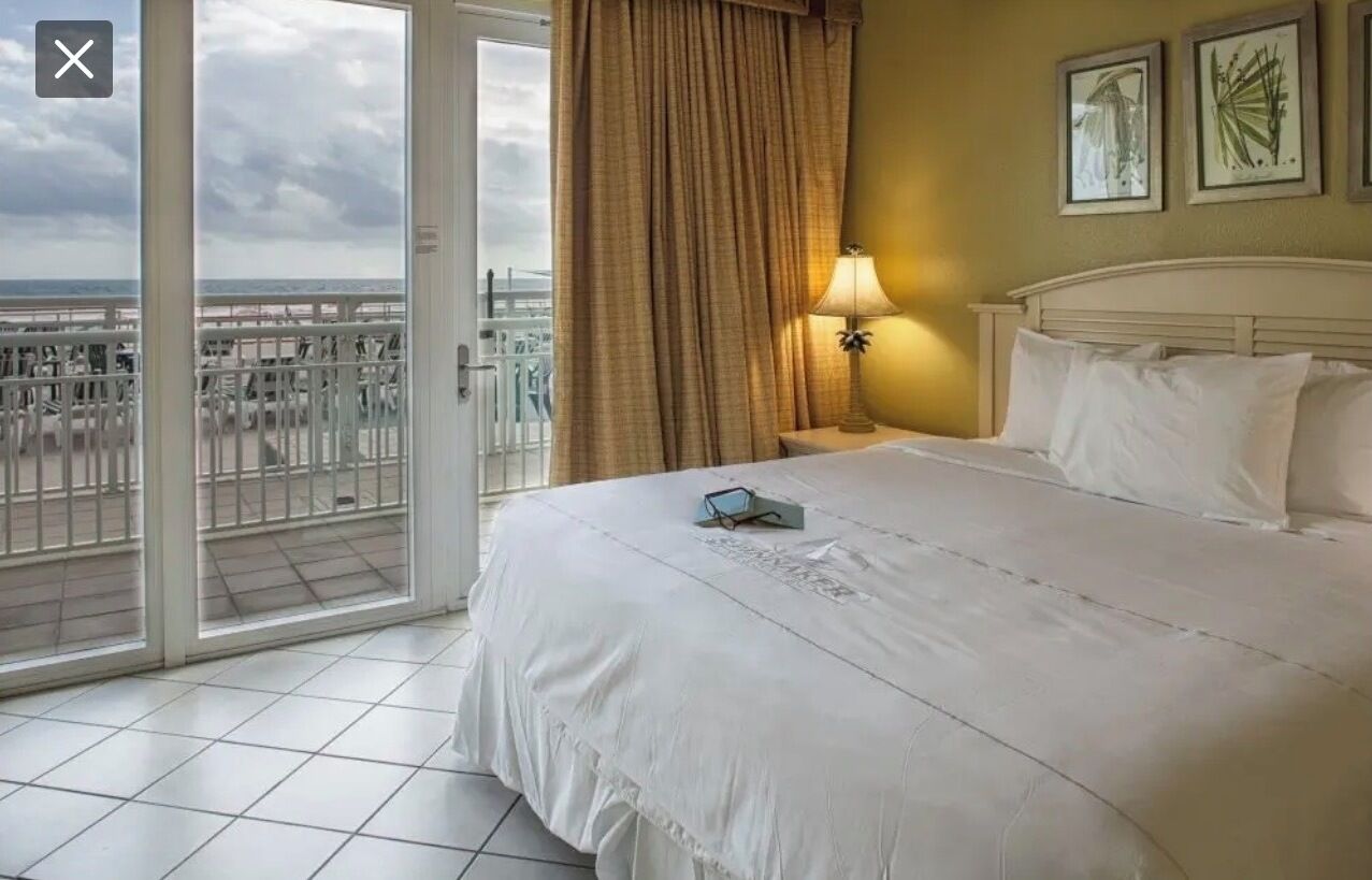 A hotel room with a bed, a nightstand, a lamp, and a view of the beach through a sliding door.