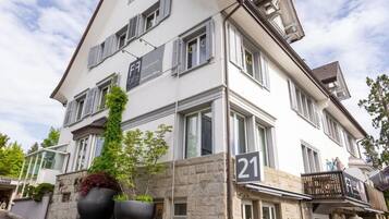 Un immeuble à plusieurs étages, de couleur blanche, portant le numéro 21, et doté d’un balcon avec des sièges pour l’extérieur.