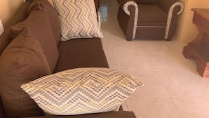 A living room with a brown sofa and armchairs, a kitchen in the background, and a dining area visible through an archway. - Charming 3-bedroom house with AC, WiFi in delightful Savanna La Mar (Savanna La Mar)