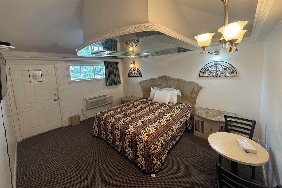 Une chambre avec un grand lit, une petite table, une chaise, un luminaire au plafond et une fenêtre.