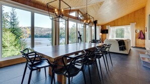 A modern kitchen with a large island, wooden ceiling, and large windows offering a view of a forest and lake. - Repère tranquille au bord du lac (Saint-Alexis-des-Monts)