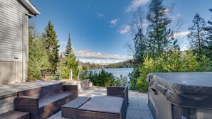 An outdoor patio with wicker furniture, a hot tub, and a view of a lake surrounded by trees. - Repère tranquille au bord du lac (Saint-Alexis-des-Monts)