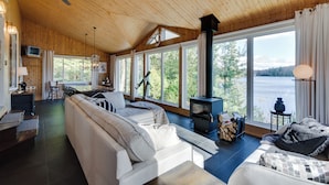 A cozy cabin interior with a large sofa, a dining area, and a wood-burning stove. - Repère tranquille au bord du lac (Saint-Alexis-des-Monts)