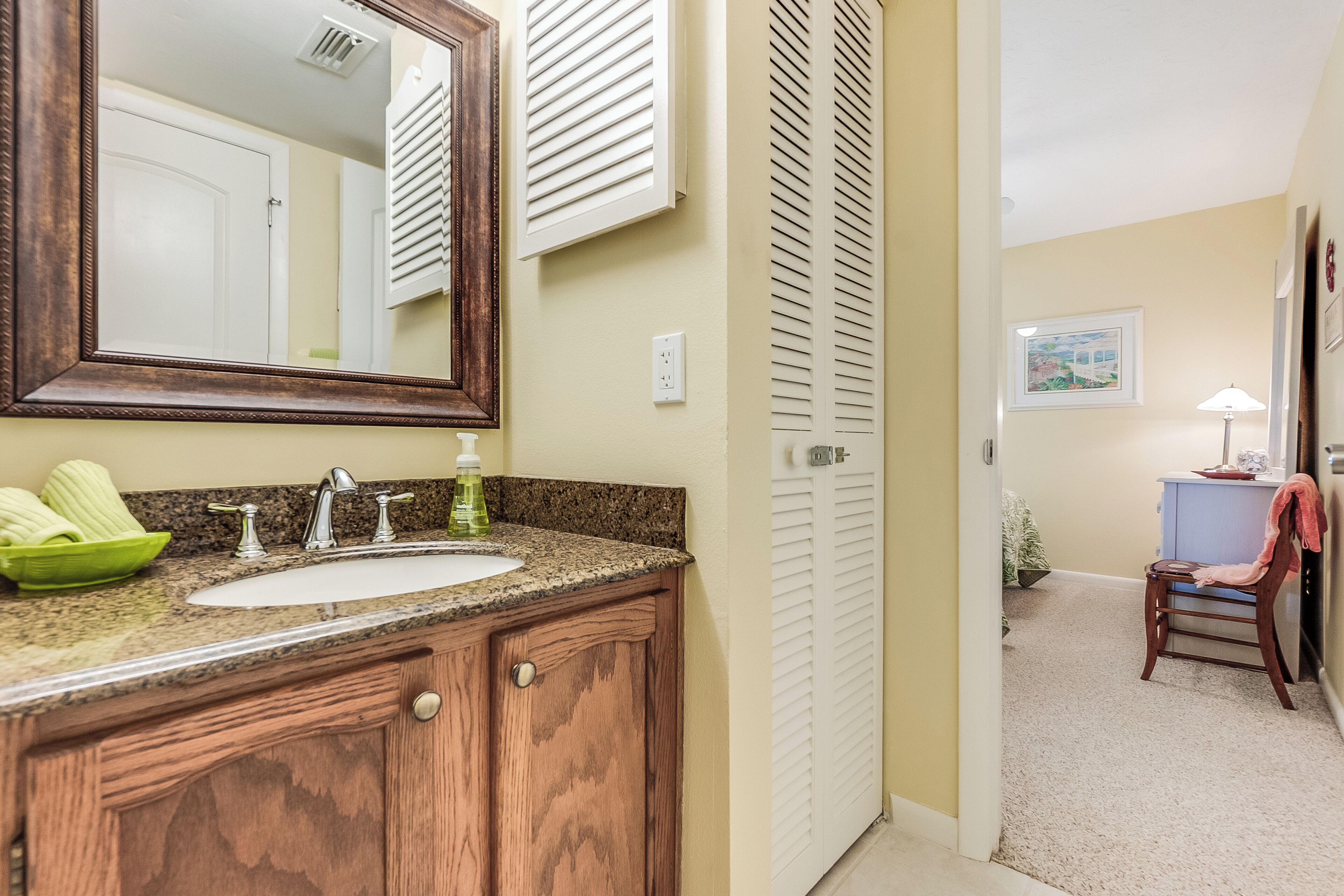 Une salle de bain avec un meuble-lavabo en bois, un évier et un miroir. On y trouve une porte avec des volets à persiennes et une petite chaise sur laquelle est posée une serviette.