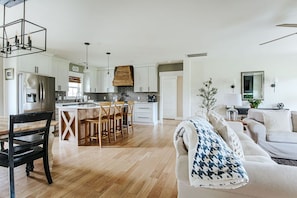 A modern kitchen with white cabinets, a dining area with wooden chairs, and a living room with a sofa and a decorative plant.