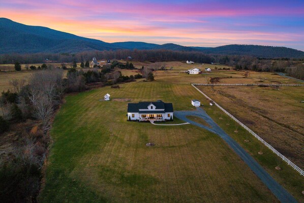 A large, single-story house with a central driveway and a fenced yard, surrounded by open fields and distant hills.