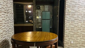 A dining area with a round wooden table and four chairs, a refrigerator, and a window with blinds.