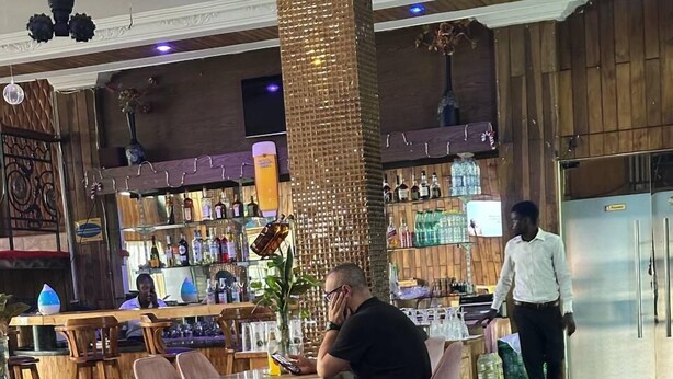 Een restaurant met een bar, zitplaatsen met blauwe stoelen, en een persoon op de achtergrond.