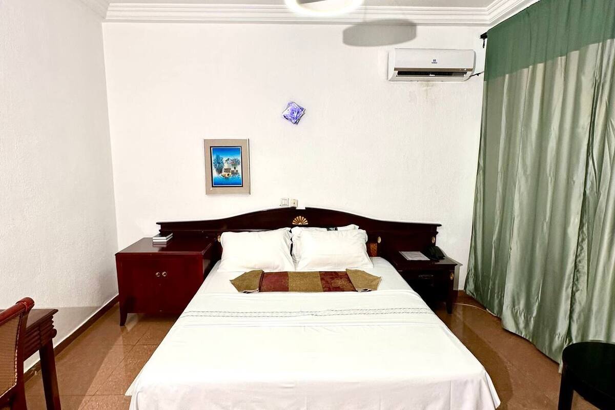 Quarto de hotel com uma cama grande, mesa de cabeceira de madeira, escrivaninha, cadeira, ar condicionado e cortina verde.
