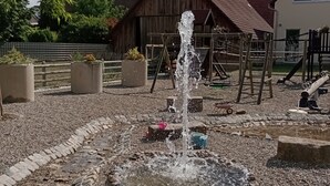 Una fuente de piedra en un patio con un parque infantil y un edificio de madera al fondo.