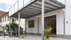 Un área de estar al aire libre techada con sillas y plantas en macetas, una entrada de edificio con puerta de cristal y un letrero con la indicación del lugar.