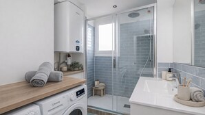 A modern bathroom with a washing machine, a towel rack, a glass shower enclosure, and a sink with toiletries.
