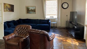 Ein Wohnzimmer mit blauem Sofa, zwei Ledersesseln, einer Uhr, einem Fernseher und einem Fenster mit Jalousien.