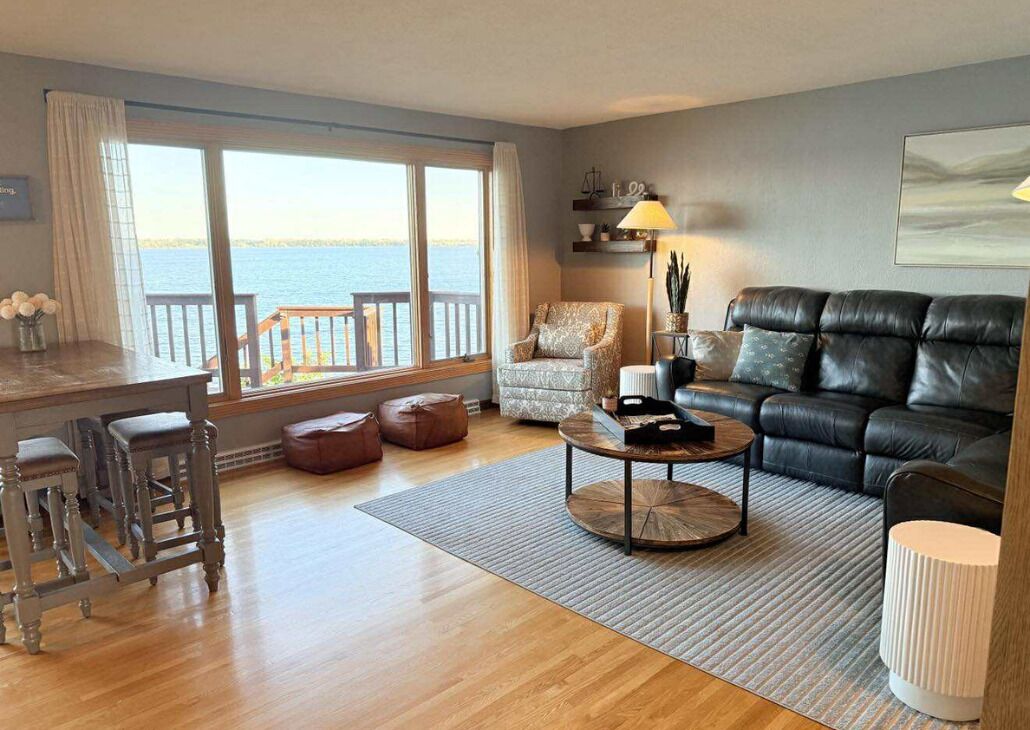 Una sala de estar con vista al mar, un sofá de cuero negro, un sillón estampado, una mesa de centro de madera y una zona de bar con taburetes.
