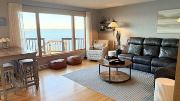 Una sala de estar con vista al mar, un sofá de cuero negro, un sillón estampado, una mesa de centro de madera y una zona de bar con taburetes.