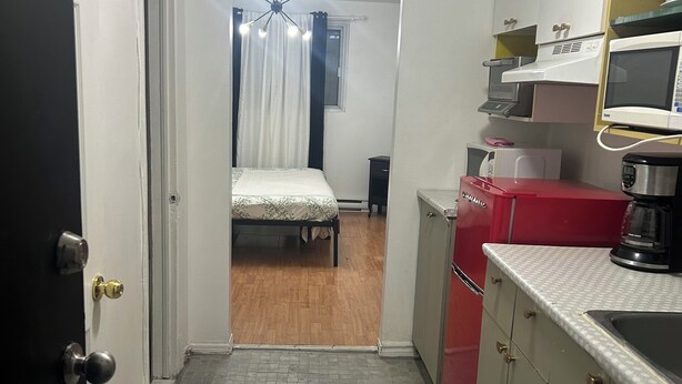 A narrow hallway leading to a bedroom with a bed and a window with curtains. There is a kitchenette with a microwave, a red appliance, and a coffee maker.