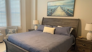 Ein Schlafzimmer mit einem großen Bett, zwei Nachttischen, einem Beistelltisch, einem Sessel und einem Gemälde eines Piers.
